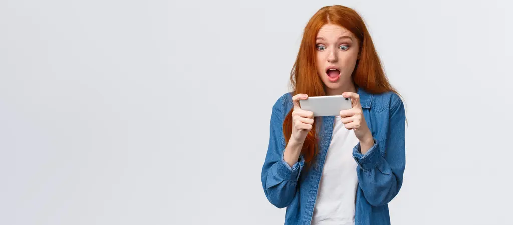 Mobiltelefonon j&aacute;tsz&oacute; felhaszn&aacute;l&oacute; meglepett arckifejez&eacute;ssel online j&aacute;t&eacute;k k&ouml;zben